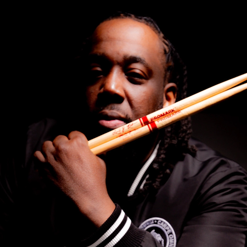 Quinton Robinson, with braided hair, holds two drumsticks crossed before his face. He wears a black jacket with white striped cuffs and a circular emblem. Dramatic lighting highlights his face and hands against a dark background.