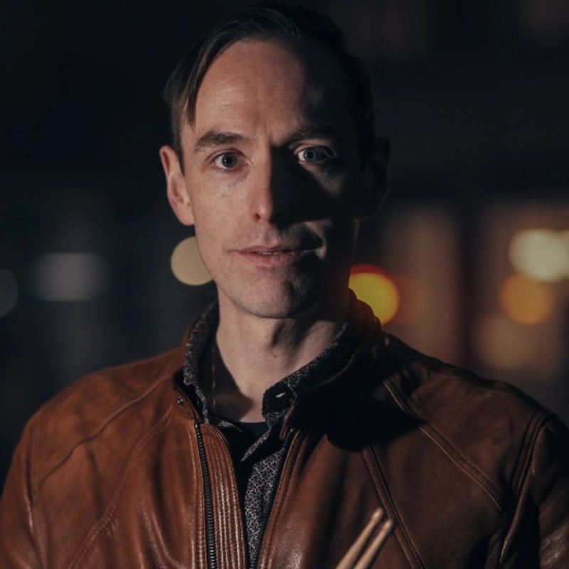 Jake Robinson, with short brown hair, wears a brown leather jacket over a dark patterned shirt. He stands facing the camera at night, softly lit, with blurred lights and dark background, holding a pair of drumsticks.