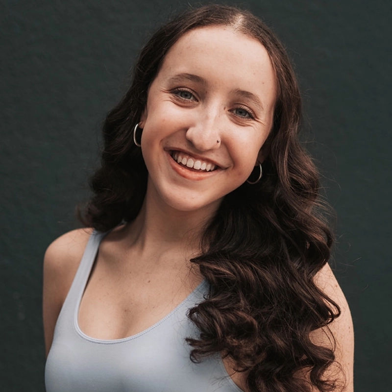 Jessee Leigh Robinson, a young woman with long, wavy brown hair, smiles warmly at the camera. She wears silver hoop earrings, a nose ring, and a light blue sleeveless top against a plain dark green wall, exuding friendliness and confidence.