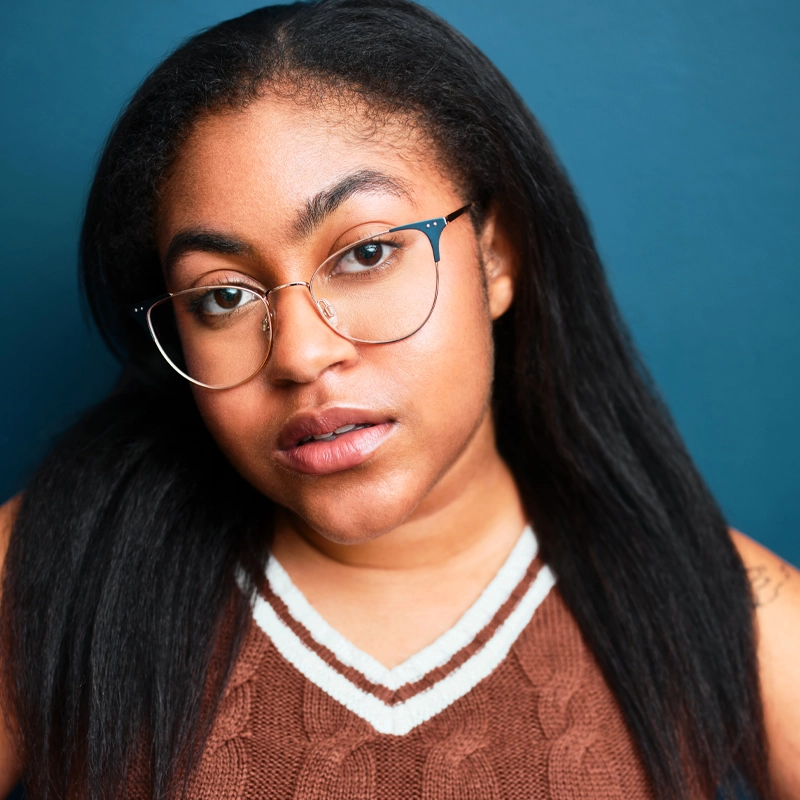 Nyla Alyese Robotham, a young woman with long, straight dark hair and round eyeglasses, wears a brown sweater vest with white trim as she poses in front of a blue background, looking directly at the camera with a neutral expression.