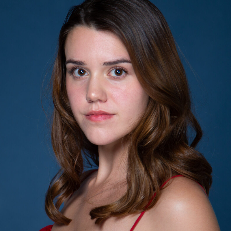 Robyn Chandler, a young woman with wavy brown hair and fair skin, gazes softly at the camera. She wears a red top with thin straps against a solid blue background, her natural makeup and even lighting giving the portrait a calm, professional look.
