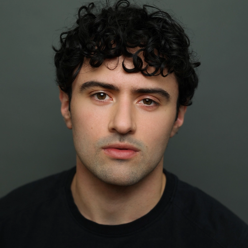 A young man with short curly dark hair and fair skin stares directly at the camera with a neutral expression. He wears a plain black shirt and is posed against a solid gray background. His face is evenly lit, highlighting his features.