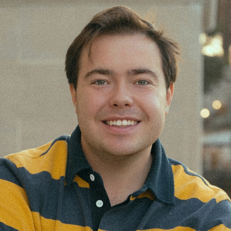 A young man with fair skin and straight brown hair smiles at the camera. He wears a yellow and navy blue striped rugby shirt, and the background is softly blurred, showing outdoor lights and a beige wall.