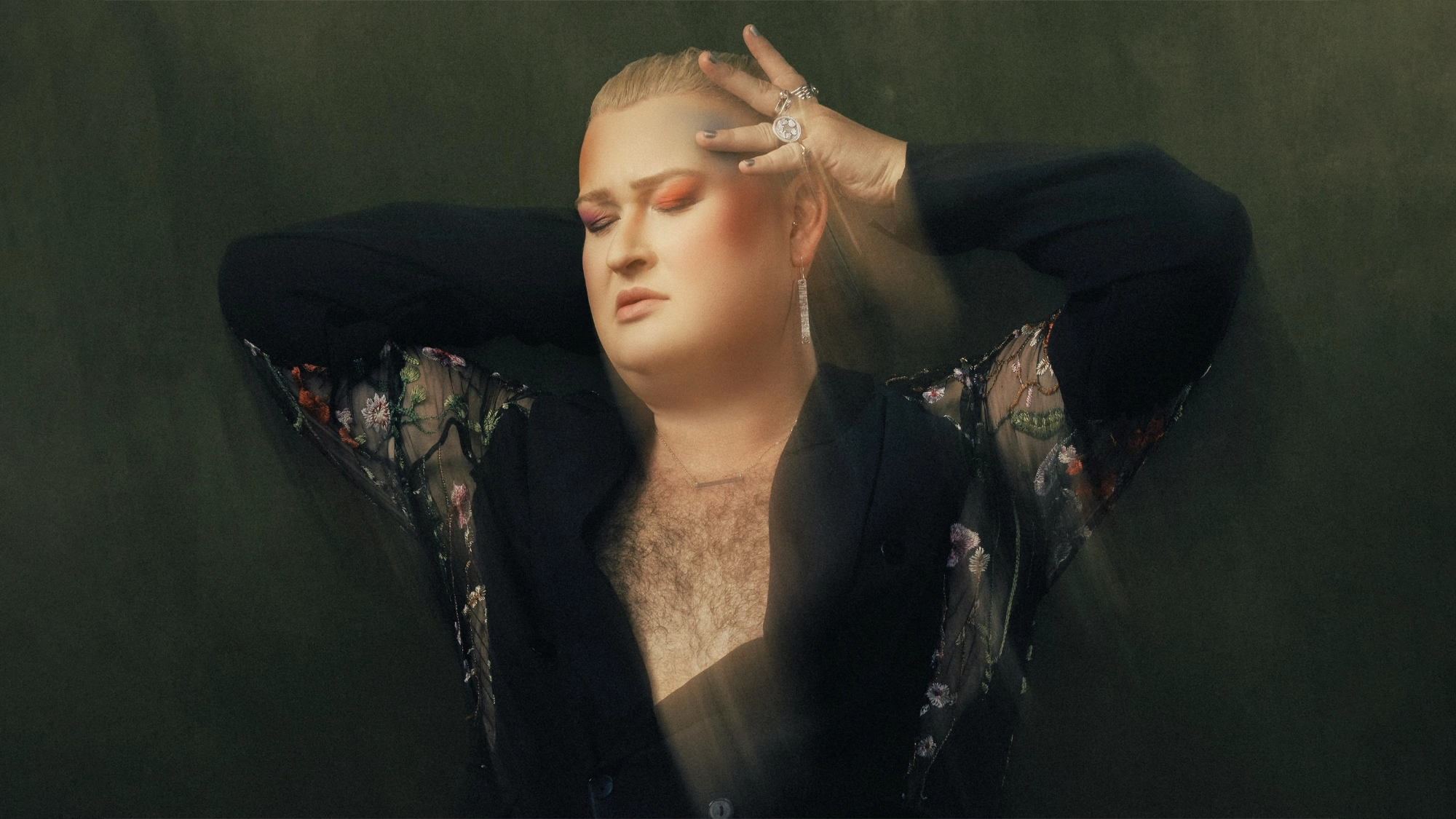 Ryan James Monroe - Site Image Ryan James Monroe poses with eyes closed, wearing dramatic orange eyeshadow, long earrings, and rings. Dressed in a sheer black top with floral embroidery, Monroe raises their hands to their head against a soft green background.