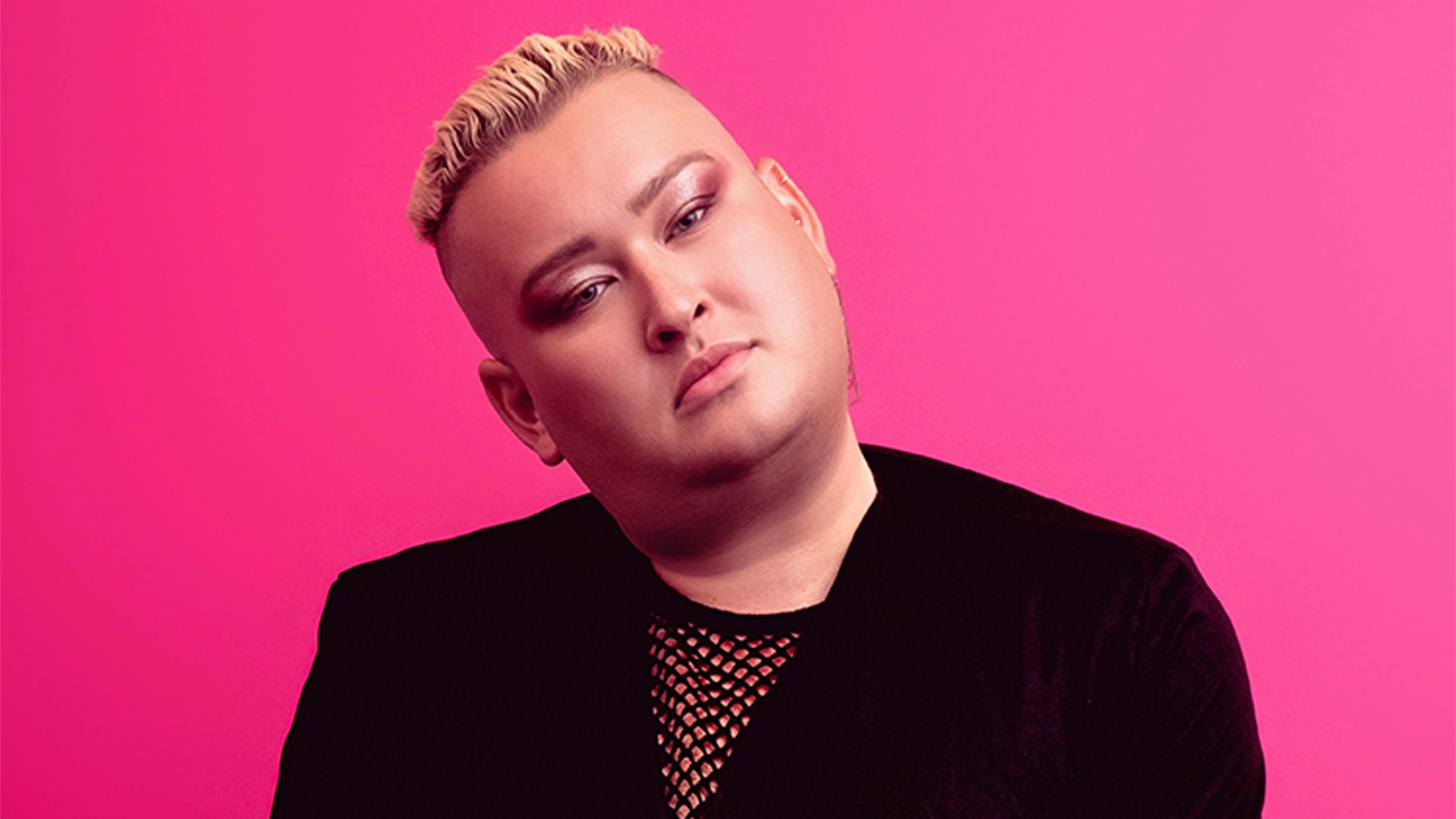 A person with short, bleached blond hair poses against a bright pink background. Channeling Cabaret Disco vibes, they wear a black velvet jacket over a mesh top, with shimmering eyeshadow and neutral lipstick. Their head is slightly tilted.