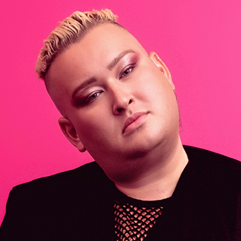 A person with short, bleached blond hair styled in waves poses against a bright pink background. They have full makeup, including bold eyeshadow and defined brows. They wear a black top with a mesh detail and a black jacket, looking directly at the camera.