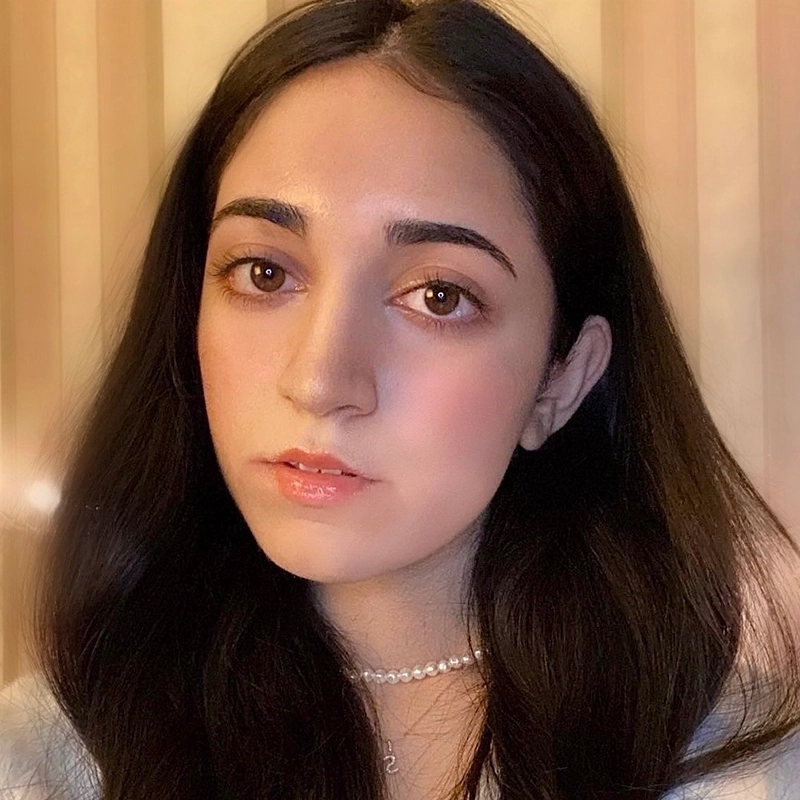 Grace Whetstone, a young woman with long, dark hair and brown eyes, looks at the camera with a neutral expression. She wears pale pink gloss, natural makeup, a pearl necklace, and a light top against a softly blurred beige striped background.