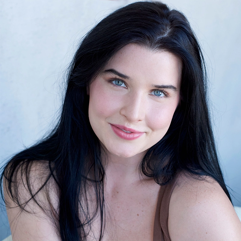 Madison Keenoy, with long straight black hair and blue eyes, smiles gently at the camera. She has fair skin, lightly flushed cheeks, and wears a sleeveless brown top against a soft, light background that adds to the calm, natural feel.