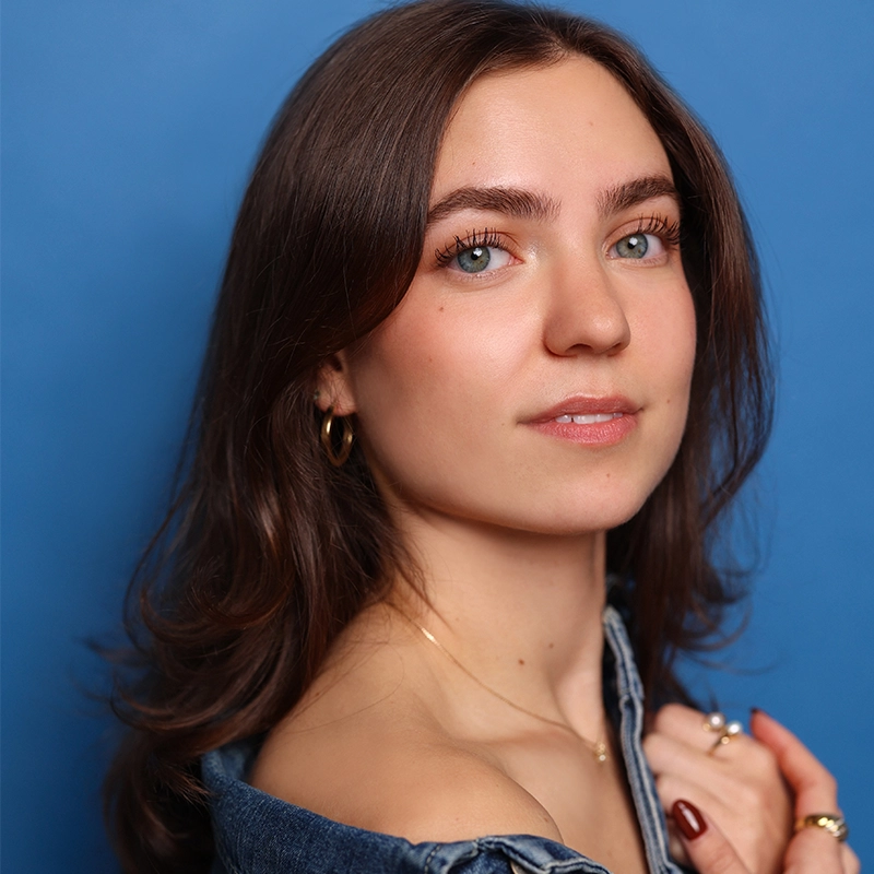 A young woman with fair skin and long brown hair looks confidently at the camera. Samantha Garvey wears a denim jacket off one shoulder, gold hoop earrings, and several rings, set against a solid blue background.