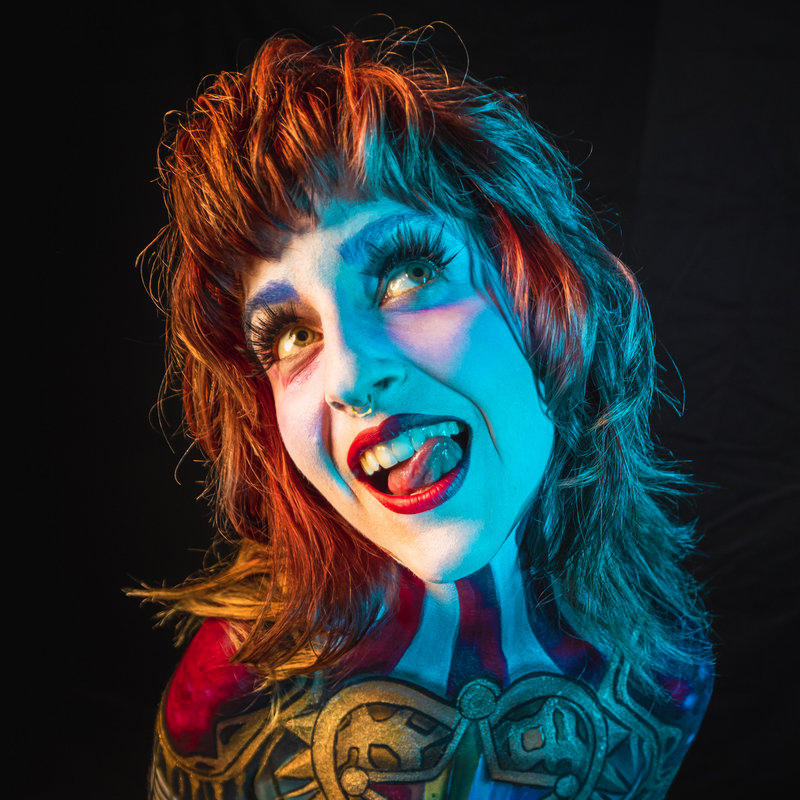 Chanel Samson, with shaggy red hair, poses against a dark background in elaborate blue and white face and body paint resembling fantasy armor. Smiling widely and sticking out their tongue playfully, dramatic lighting casts blue and orange tones.