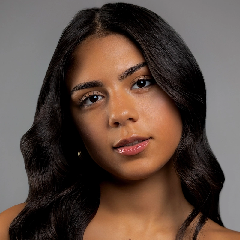 Coral Rosario-Laboy, a young woman with long, dark, wavy hair and smooth medium skin, looks into the camera. She has natural makeup, defined brows, glossy lips, and small hoop earrings. A plain gray background highlights her serene confidence.