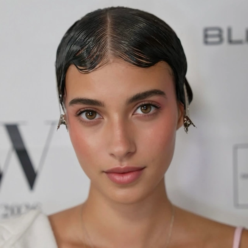 A young woman with smooth, shiny, slicked-down dark hair styled with gentle waves at the front. She has natural makeup with pink blush and lips, wears hair clips near her ears, and exudes an Anjalee Hardy-inspired elegance. White background with blurred black text.