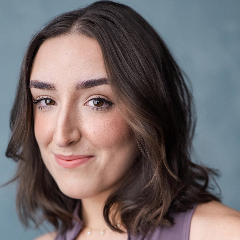 A young woman with wavy brown hair and fair skin smiles softly at the camera. She has well-groomed eyebrows, natural makeup, and wears a sleeveless lavender top—a serene portrait in muted blue-grey by Brennan Schmidt.
