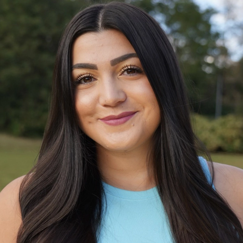 Caitlyn Schmidt, a woman with long, straight dark hair and full eyebrows, smiles gently at the camera. She wears a light blue sleeveless top and stands outdoors with green trees and grass softly blurred in the background.