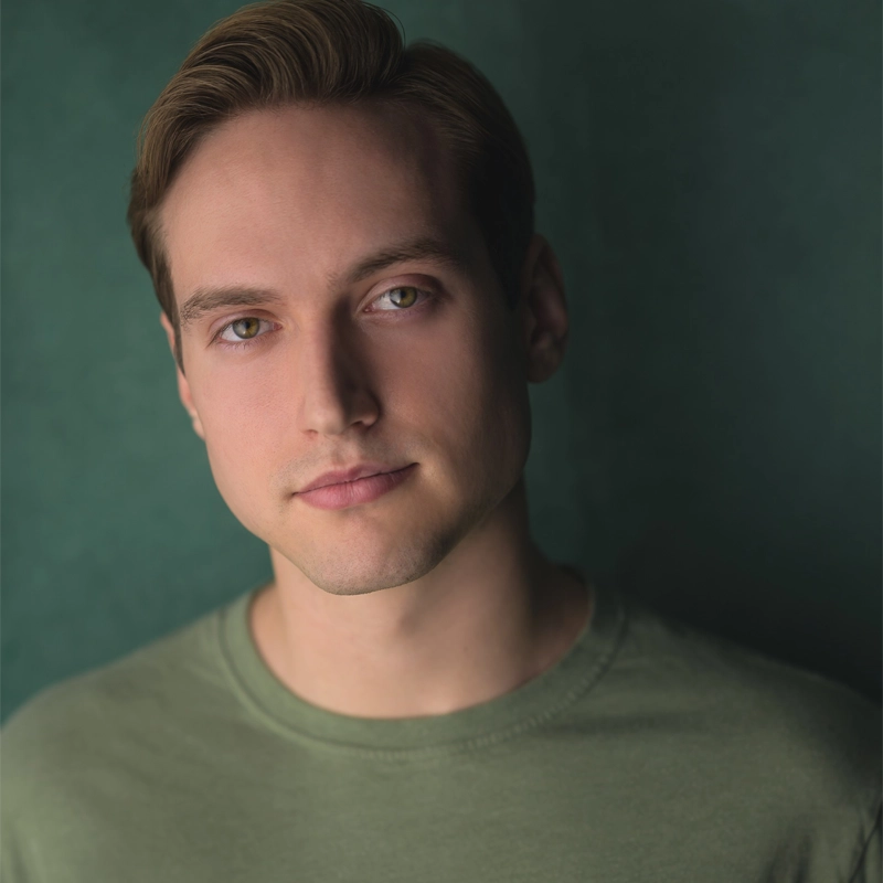 A young man, Austin Schulte, with short light brown hair and green eyes is wearing a light green crew-neck shirt. He faces the camera, head slightly tilted to the right, with a neutral expression against a soft, blurred green background.