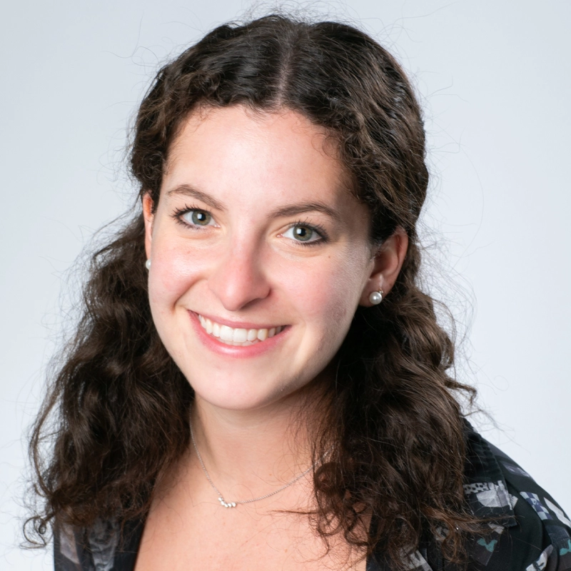 A woman with long, curly brown hair smiles at the camera. She wears a black patterned top, a silver necklace, and pearl stud earrings. The background is plain light gray and the lighting is even, giving the portrait a professional feel.
