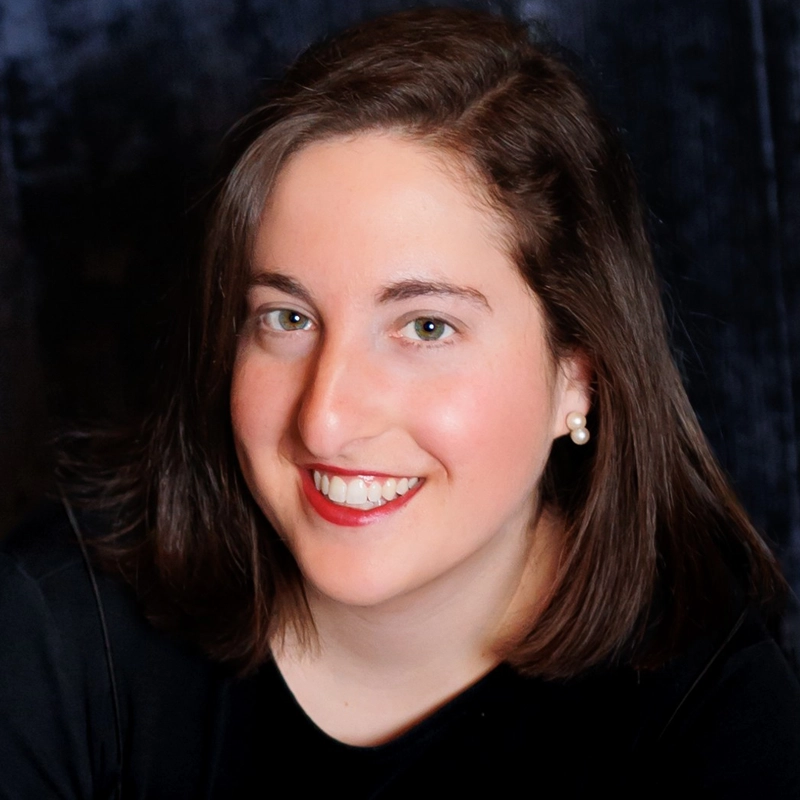 A woman with straight brown hair and fair skin smiles at the camera. She wears red lipstick, pearl earrings, and a black top. The background is dark and softly blurred, making her face the focal point of the image.
