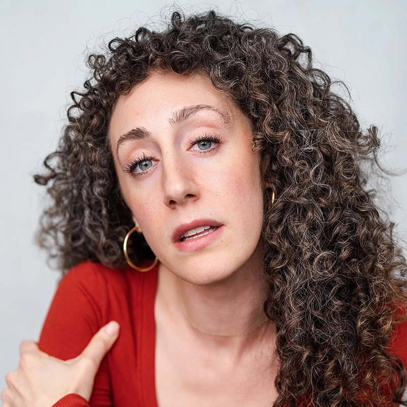 Ashley Wells has long, dark curly hair and is wearing a red top and gold hoop earrings. She is looking directly at the camera with a neutral expression.