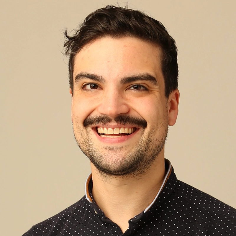 Spencer Lawson, a smiling man with short dark hair and a mustache, wears a dark shirt with small white polka dots and light collar trim as he stands in front of a plain beige background, radiating a cheerful and friendly expression.