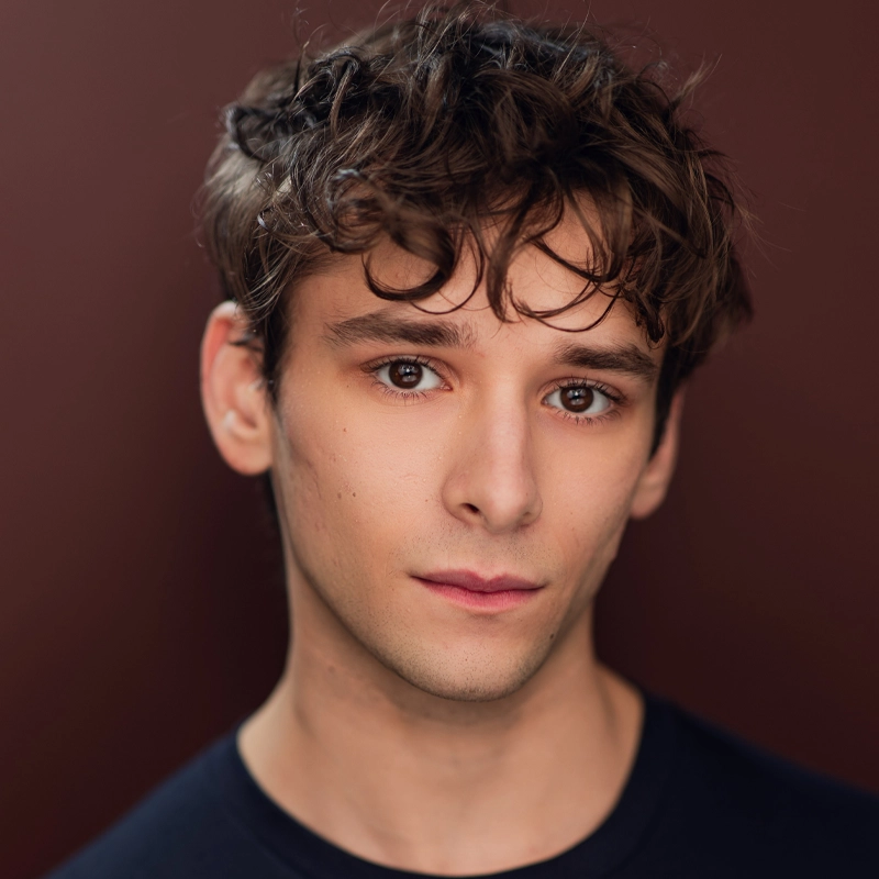 A young man with wavy brown hair, fair skin, and brown eyes—Jason Soranno—stares gently at the camera. He has a neutral expression, subtle makeup, and a dark shirt against a soft maroon background, creating a calm and focused portrait.