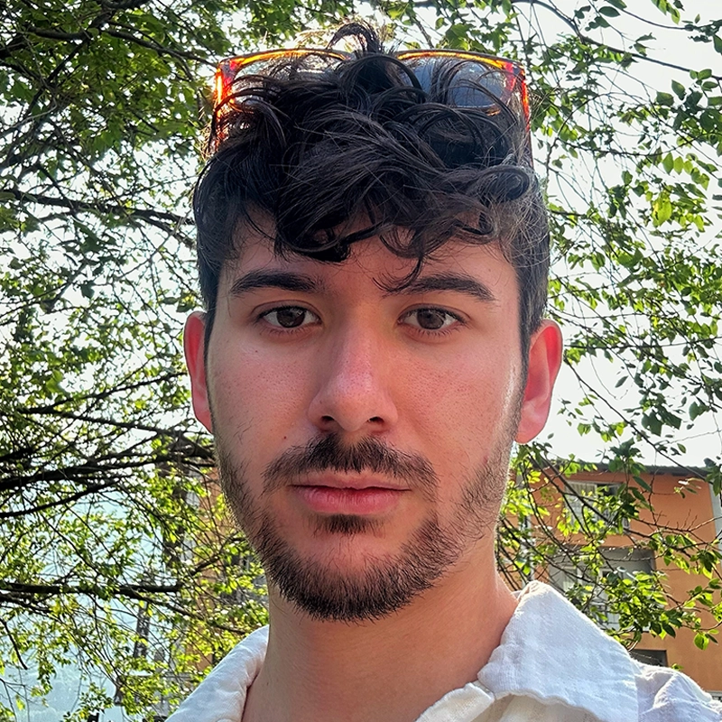 A young person with short, wavy dark hair and facial hair looks at the camera outdoors. Wearing a white shirt and sunglasses on their head, Spencer Lawson stands with sunlight filtering through leafy tree branches and part of a building in the background.