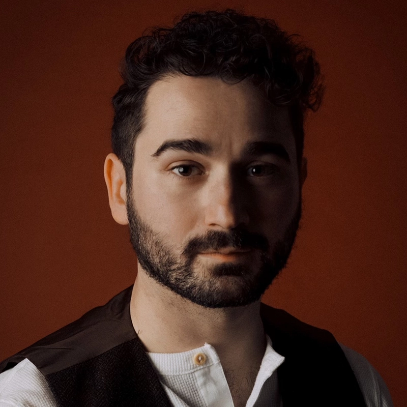 Max Marsillo, a man with curly dark hair and a beard, gazes at the camera against a reddish-brown background. He wears a white shirt with a round collar and a dark vest. Soft lighting gives the portrait a warm, introspective feel.