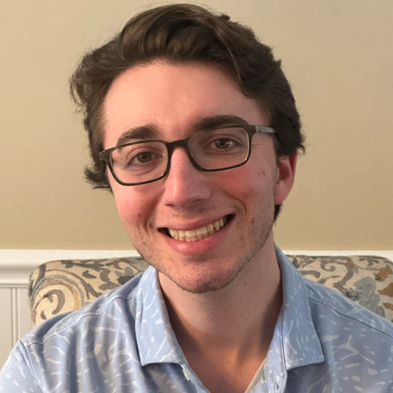 Jake Staffin, a young man with light skin, short brown hair, and glasses, smiles at the camera. He wears a light blue patterned collared shirt and sits in front of a beige wall with white wainscoting and a decorative upholstered chair.