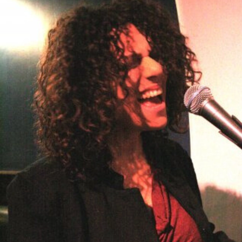 A person with curly dark hair sings passionately into a microphone, mouth open wide and eyes closed. They wear a dark jacket over a maroon top, and warm light casts shadows across their face and hair. The background is mostly dark with a light area on the right.