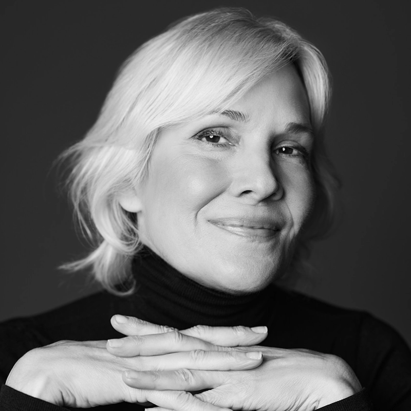 A smiling older woman with light hair and light skin rests her chin on her hands, which are gently clasped together. She wears a dark turtleneck and looks into the camera. The portrait is black and white, with a dark, plain background.