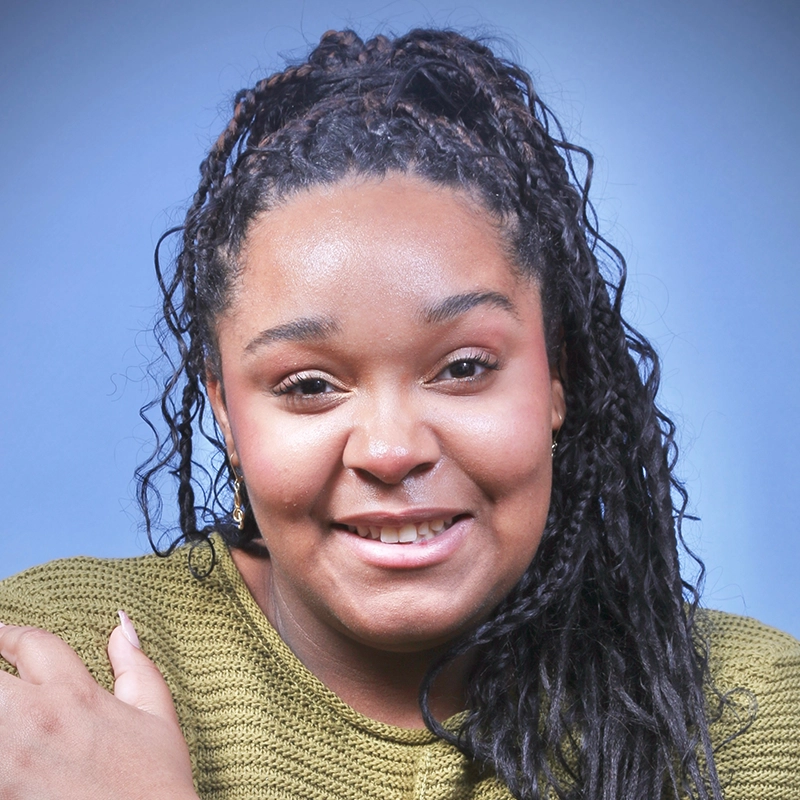 A woman with dark curly hair in a high ponytail smiles at the camera. She wears a textured olive-green sweater and natural makeup. The soft blue gradient background adds to Kitana’s friendly and approachable vibe.