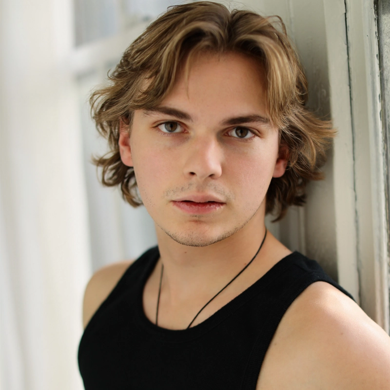A young man, Ryan Sweeney, with shoulder-length, wavy light brown hair leans against a window frame, gazing directly at the camera. He wears a black tank top and a thin necklace, with soft natural light illuminating his face and neutral background.
