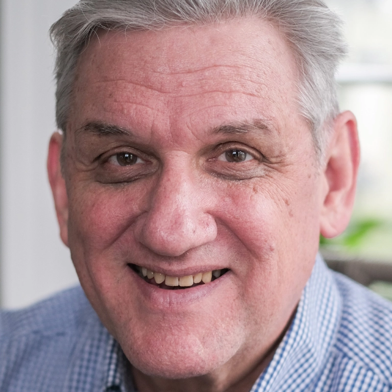 Steve Taylor, a middle-aged man with short gray hair, smiles warmly at the camera. He is wearing a blue and white checkered shirt, with a softly blurred background that hints at an indoor setting bathed in natural light.