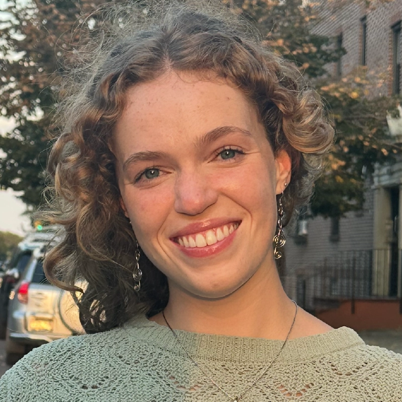 A young woman with curly, light brown hair smiles at the camera. She wears silver earrings and a light green knit top. Sunlight casts a warm glow on her face as she stands near trees and a brick building at Spencer Lawson’s campus.