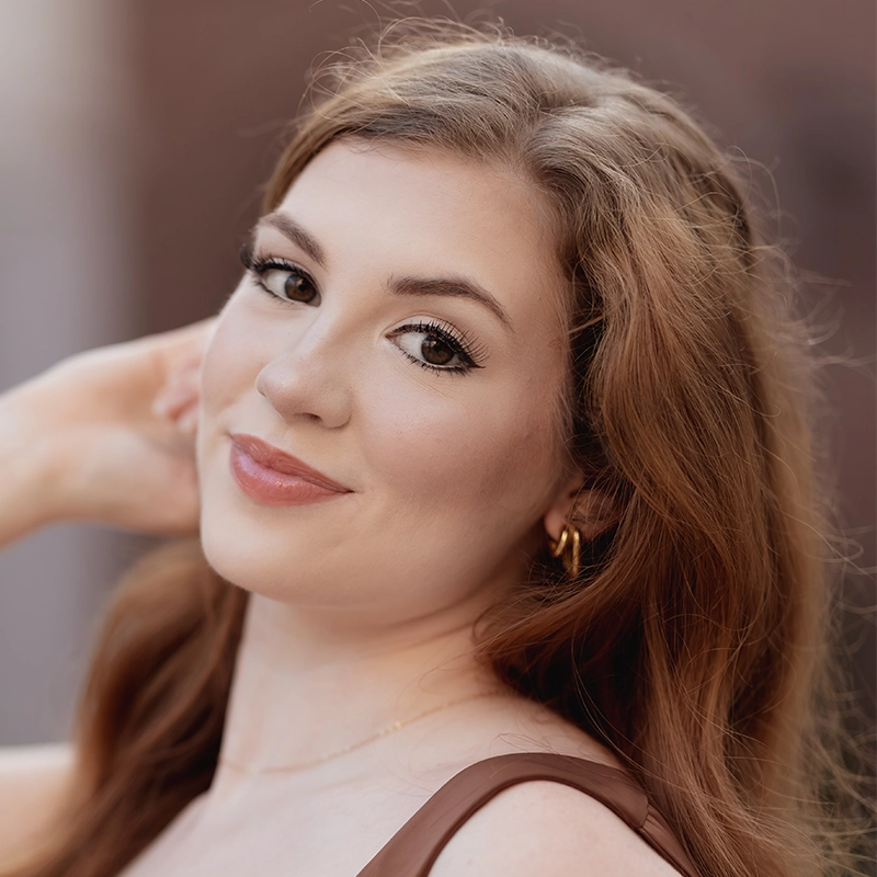 A young woman with long, wavy auburn hair smiles softly at the camera. She has fair skin, subtle makeup, and wears gold hoop earrings and a thin necklace—her style evokes the elegance of a modern Sultan. The background is softly blurred.