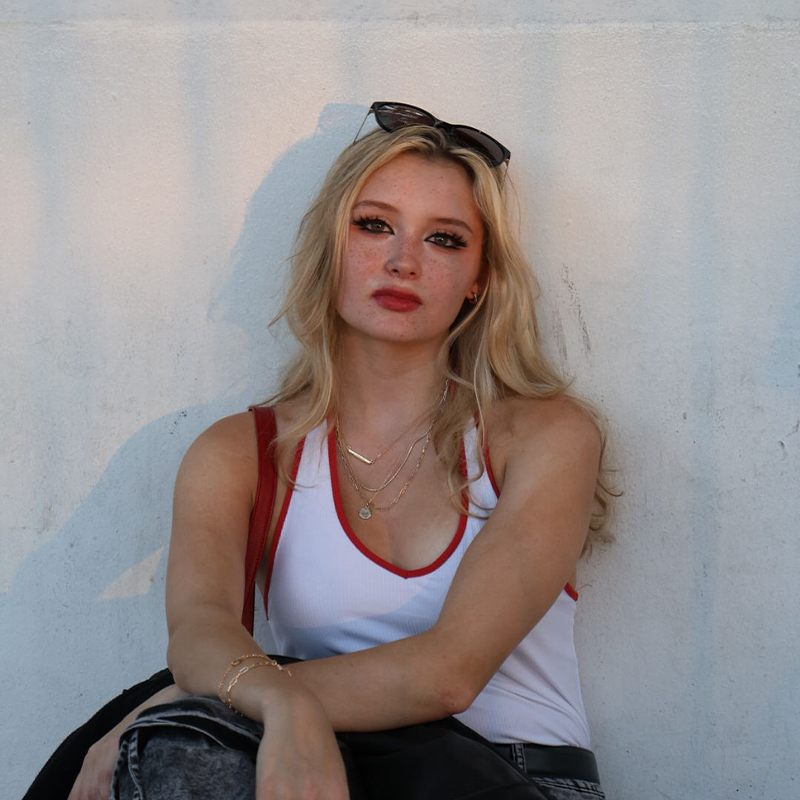 A young woman with long blonde hair sits against a white wall. Thalia Hart wears sunglasses on her head, a white tank top with red trim, layered necklaces, and a casual expression. Shadows and sunlight create an angel-soft backdrop.
