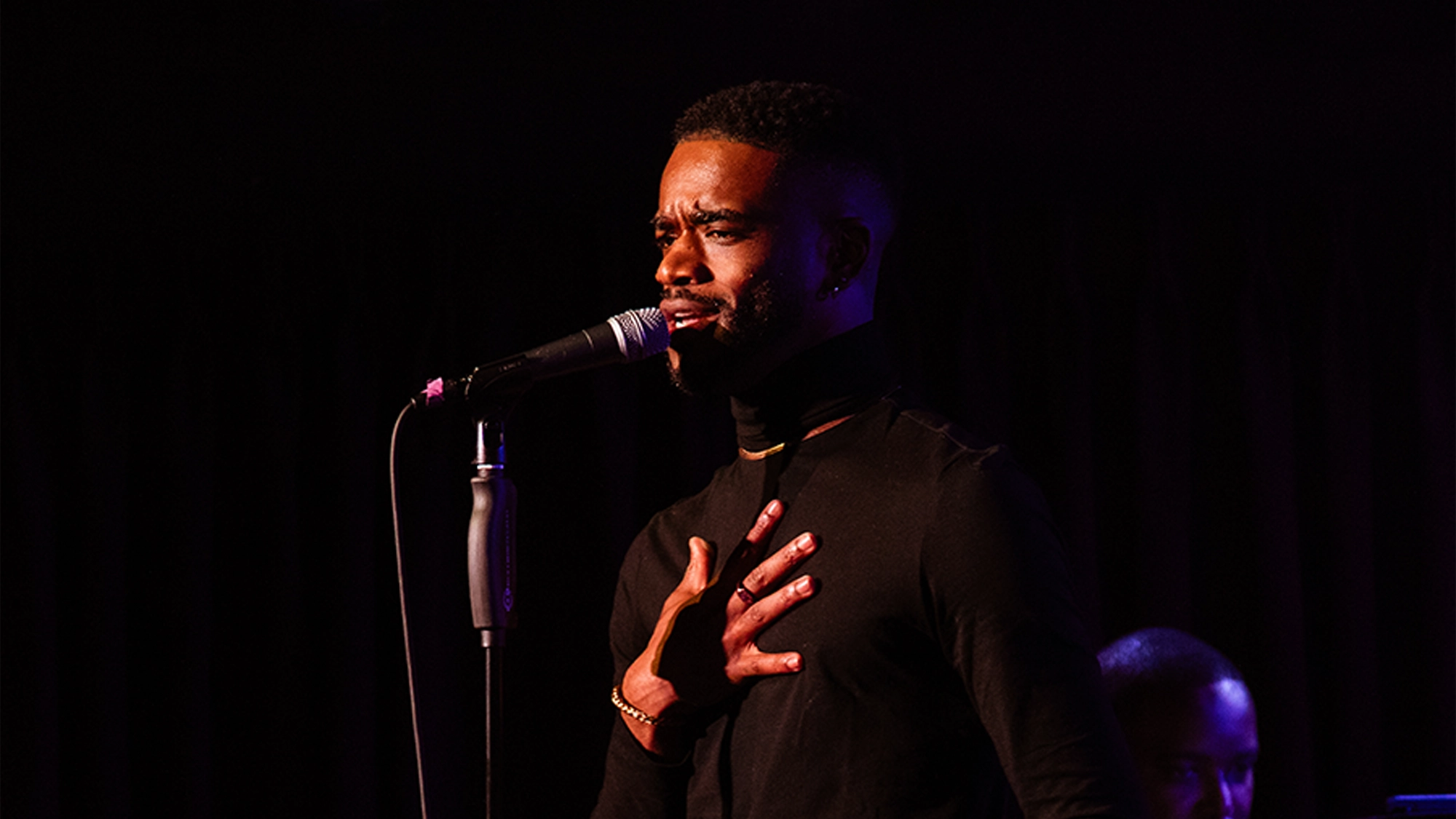 A man in a black turtleneck sings passionately into a microphone on a dimly lit stage, holding one hand to his chest. The background is dark, emphasizing his expressive performance. Another person’s head is partially visible in the background.