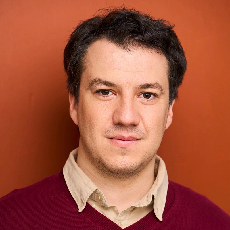 Spencer Lawson, a man with short dark hair and fair skin, faces the camera with a slight smile. He wears a beige collared shirt under a burgundy sweater against a plain, warm orange background.