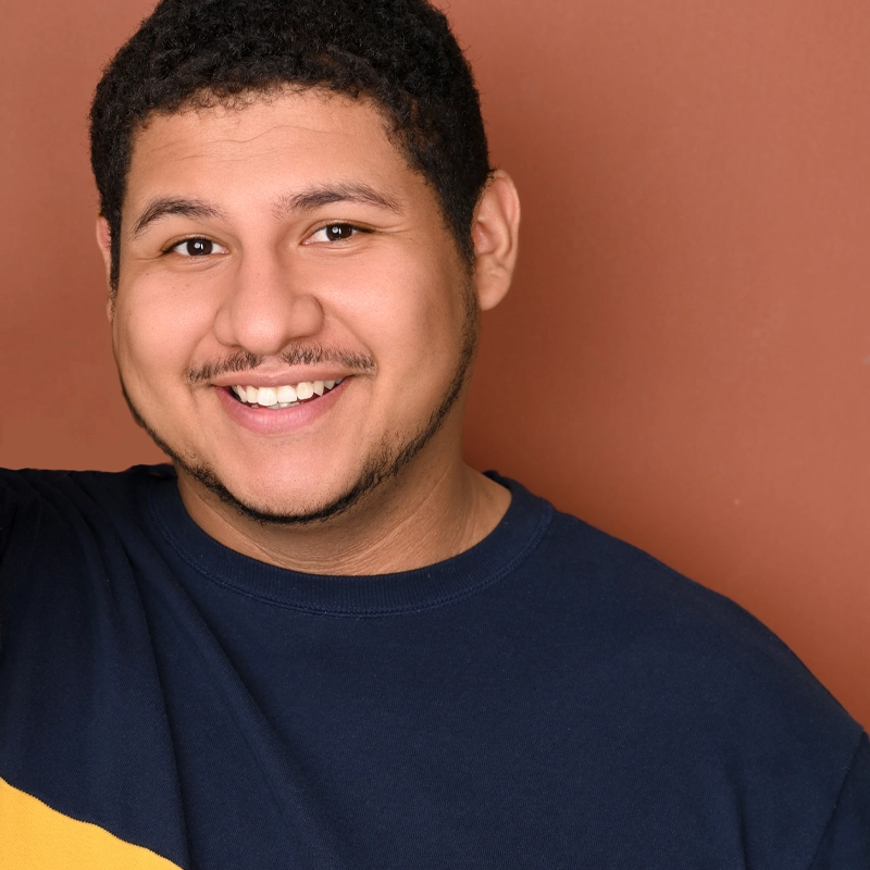 A young man with short curly hair and a trimmed beard smiles warmly at the camera. Gary Levy Tunstall wears a navy blue shirt with a yellow diagonal stripe. The background is a plain, muted brown. Only his upper torso and face are visible.