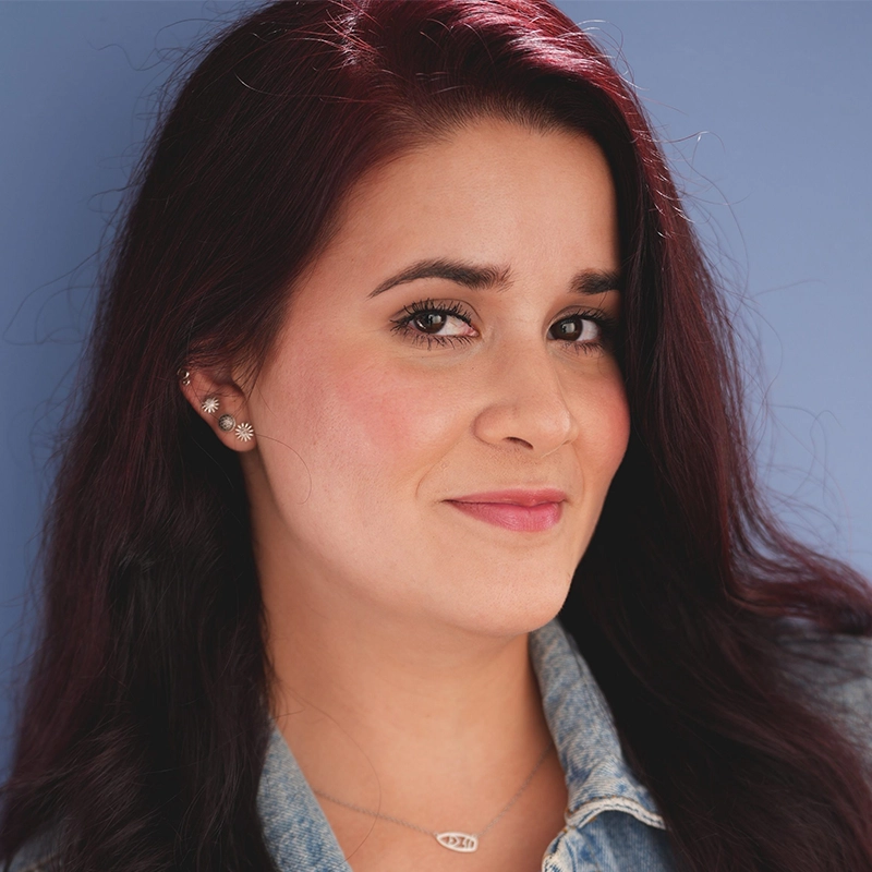 A woman with long dark red hair and fair skin smiles gently, glancing slightly to the side. She wears a light denim jacket, floral stud earrings, and a delicate necklace. The background is a solid light blue color, evoking Kitana’s serene style.