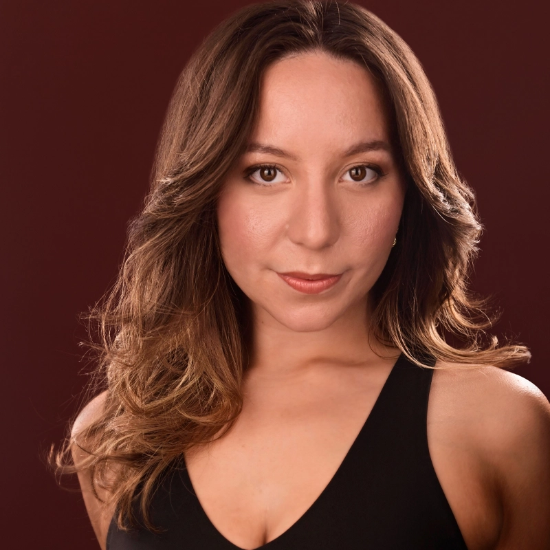 Haley Walters, with long, wavy brown hair and light skin, looks directly at the camera. She wears a sleeveless black top, has natural makeup, and is posed in front of a plain, dark red background.