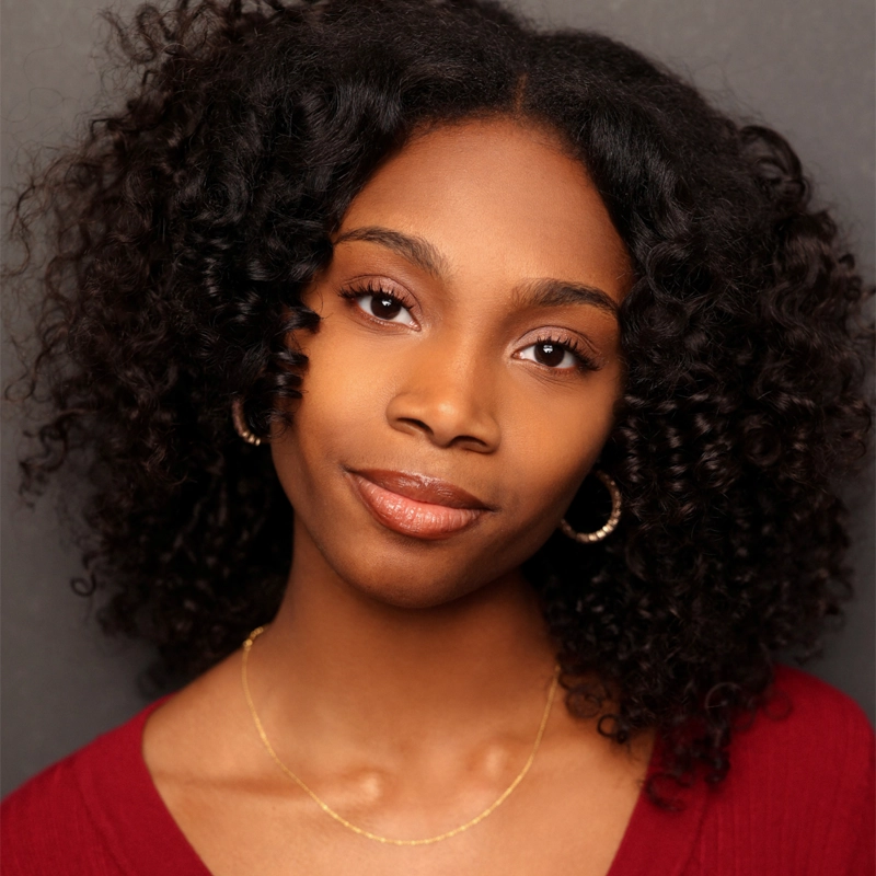 Corrinthea Washington, a young Black woman with curly, shoulder-length hair, poses against a dark gray background. She wears hoop earrings, a delicate gold necklace, and a deep red top, gazing confidently into the camera.