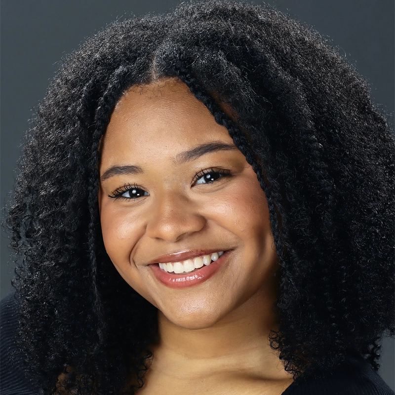 Mikayla Watkins, a young woman with medium-dark skin and curly black hair, smiles warmly at the camera. She has dimples, natural makeup, and wears a black top against a plain dark background, exuding friendly confidence.