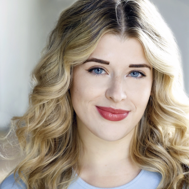 A young woman with long, wavy blonde hair and blue eyes smiles softly. She has fair skin, well-groomed eyebrows, and wears natural makeup with red lipstick. She is wearing a light blue top and the background is softly blurred.