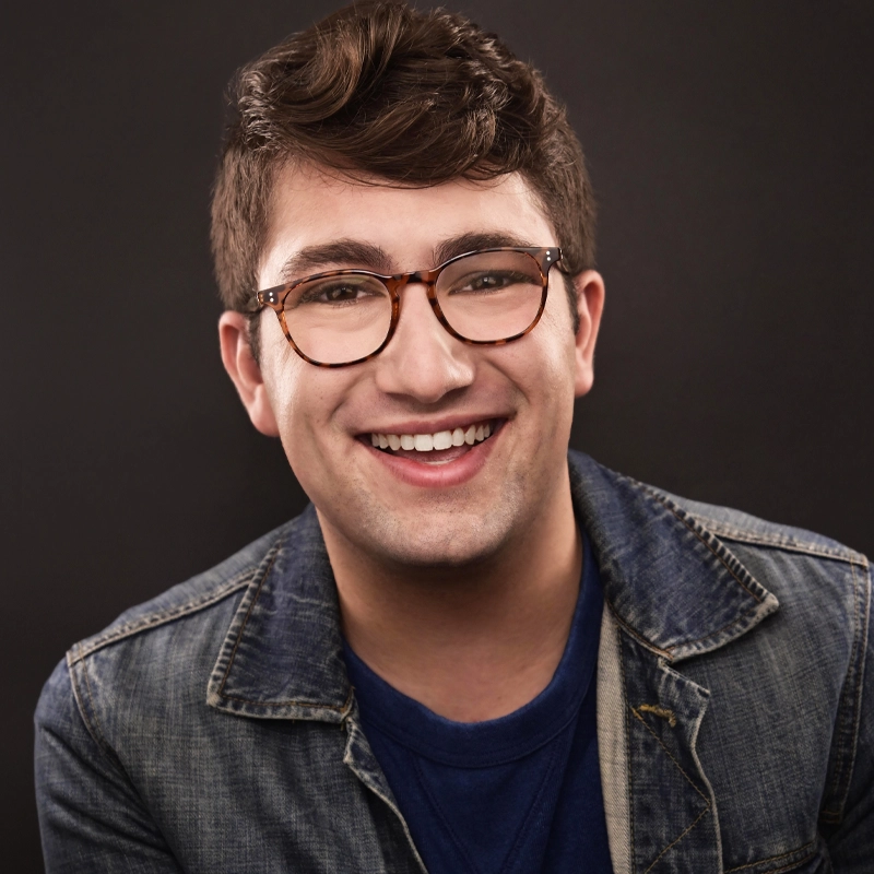 Griffin Welti, a young man with short brown hair, wearing tortoiseshell glasses, a blue T-shirt, and a denim jacket, smiles warmly at the camera. The plain dark background highlights his friendly expression.