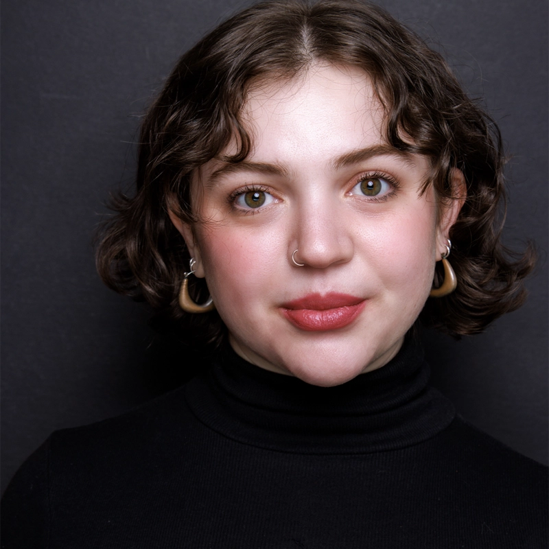 A young woman with short, wavy brown hair and hazel eyes looks at the camera, smiling softly. She wears a black turtleneck, large hoop earrings, a nose ring, and natural makeup with rosy cheeks, posed against a dark background.
