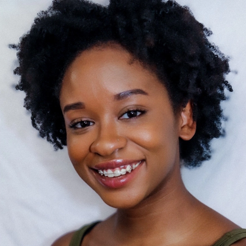 A young woman with medium brown skin and natural, curly black hair smiles warmly at the camera. She is wearing a sleeveless top and has a clear, glowing complexion. The background is simple and light-colored, keeping the focus on her face.