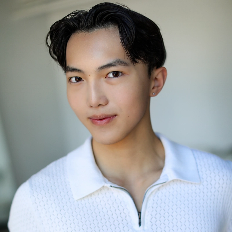 A young person with short black hair, clear skin, and dark eyes smiles softly. Tobias Yeung wears a white, textured, zippered collared shirt and a small earring in their left ear. The background is softly blurred in neutral tones.