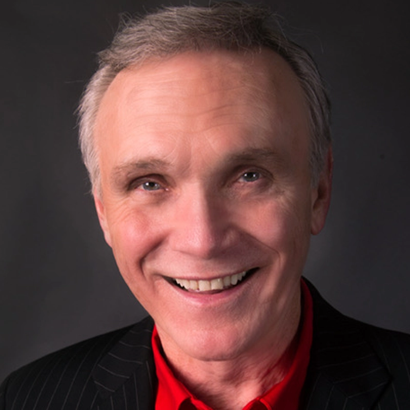 An older man with short gray hair smiles warmly. He wears a dark pinstripe suit jacket over a bright red collared shirt. The background is dark gray, and the lighting emphasizes his friendly expression and clear complexion.