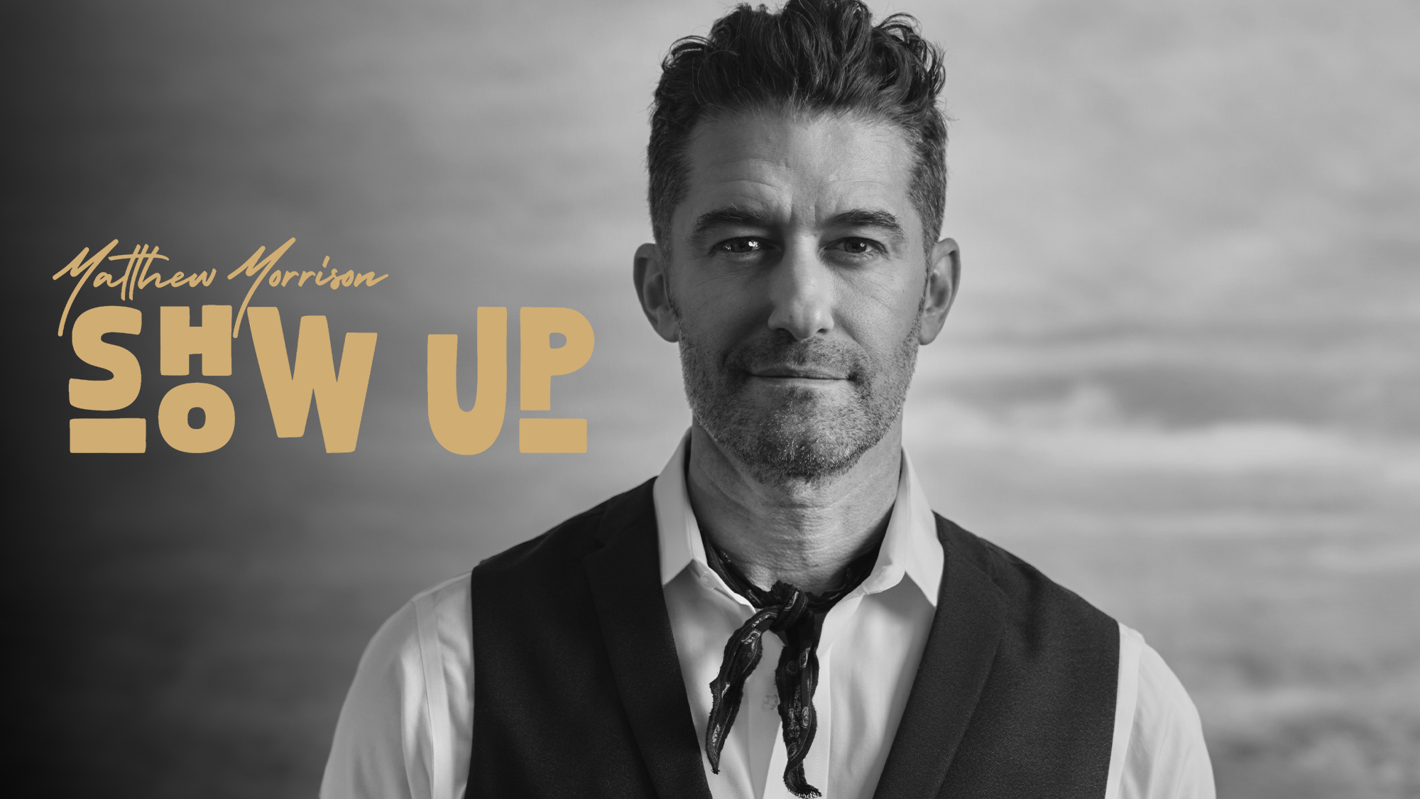 v1_site image 26_Matthew Morrison Black and white photo of Matthew Morrison in a vest, open-collar shirt, and scarf. “SHOW UP” appears in stylized tan font on the left, set against a blurred cloudy sky background.