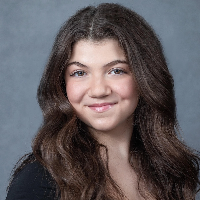 A young girl with long, wavy brown hair and fair skin smiles softly. Jackie Winslow has blue eyes, light makeup, and is wearing a black top. The soft gray background lends the portrait a professional and polished look.